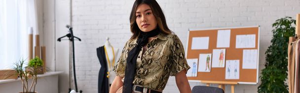 Kendine güvenen modacı Asyalı stilist kameraya özel kıyafet atölyesi, yatay afiş ile bakıyor - Fotoğraf, Görsel