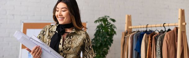 χαρούμενη ασιατική ρούχα σχεδιαστής κρατώντας μοτίβα ραπτικής στο δικό ατελιέ, επιχείρηση μόδας, πανό - Φωτογραφία, εικόνα