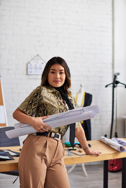 sonriente asiático ropa diseñador con costura patrones mirando cámara cerca trabajo escritorio en propio atelier - Foto, imagen