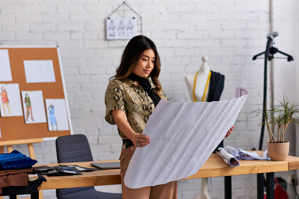 thoughtful asian fashion designer looking at sewing patterns near work desk in modern studio - Photo, Image