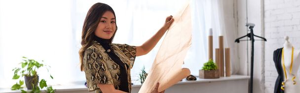 freudige kreative asiatische Stylistin mit Schnittmustern, die in ihr persönliches Atelier blicken, Banner - Foto, Bild