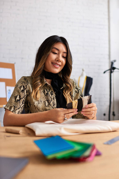 glücklich asiatische Schneiderin hält Qualitätsfäden in der Nähe von Stoff und Nähwaren in persönlichem Atelier - Foto, Bild