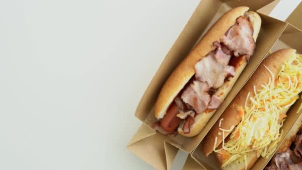 Finom hot dog, különböző feltétekkel, fehér háttérrel. Szaftos gyorskaják dobozokban - felülnézetből. Házi készítésű hotdog káposztával, szalonnával, hagymával, kolbásszal, gombával, mustárral és ketchuppal - Felvétel, videó