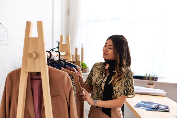 joven creativo asiático diseñador mirando a la moda a medida prendas en rack en personal atelier - Foto, Imagen