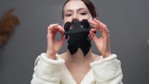 Mujer joven atractiva está quitando la máscara de sábana purificante de su cara mirando a la cámara. Concepto de cuidado de la piel facial - Metraje, vídeo