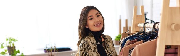 счастливый азиатский персональный стилист смотрит в камеру возле стойки с модной одеждой в ателье, баннер - Фото, изображение