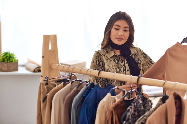 ambitious asian stylist standing near trendy casual clothes on rack in modern fashion studio - Photo, Image