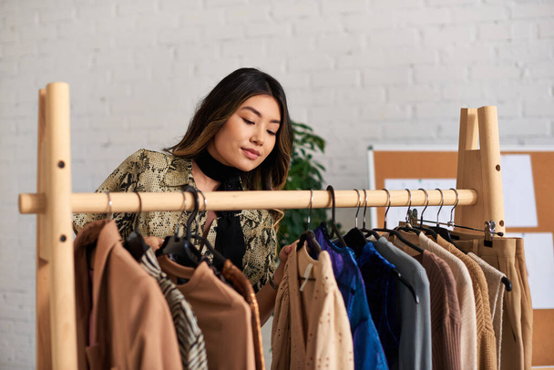 молодой и талантливый азиатский стилист смотрит на модную одежду на заказ на стойке в частном ателье - Фото, изображение