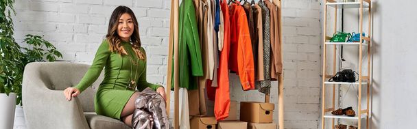 alegre elegante asiático designer em poltrona perto de rack com roupas sob medida no atelier de moda, banner - Foto, Imagem