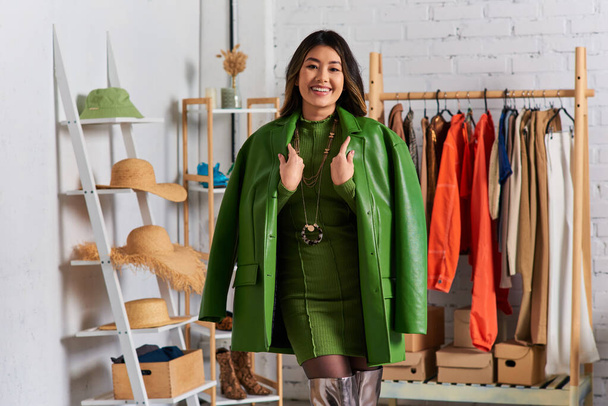 vidám ázsiai személyes stylist pózol divatos ruhák és zöld bőr kabát divat stúdió - Fotó, kép
