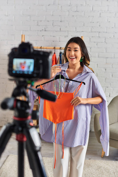 весёлый азиатский стилист, держащий топ и аксессуары рядом с цифровой камерой во время видео моды - Фото, изображение