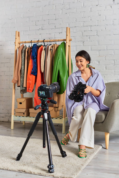 fröhliche asiatische Modedesignerin mit trendigen Sandalen während eines Videoblogs im privaten Kleideratelier - Foto, Bild