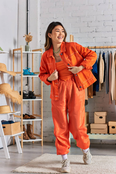 jonge vrolijke aziatische stylist in levendige casual kleding poseren in persoonlijk atelier, mode-industrie - Foto, afbeelding