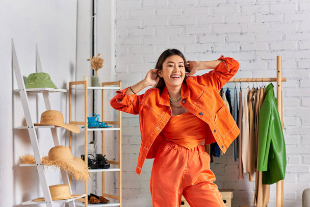 overjoyed asiatico abiti designer in luminoso arancio casual abbigliamento guardando fotocamera in proprio studio - Foto, immagini