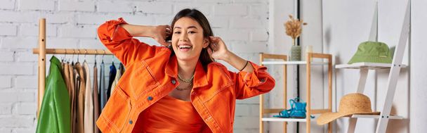 zachwycony azjatycki projektant mody pozowanie w jasnopomarańczowe ubrania w studio, baner poziomy - Zdjęcie, obraz