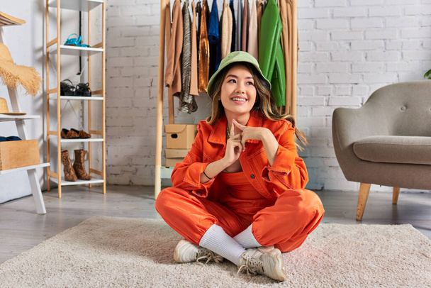 δημιουργικός ασιάτης σχεδιαστής σε panama καπέλο και πορτοκαλί ρούχα κάθεται στο πάτωμα σε ιδιωτικό στούντιο μόδας - Φωτογραφία, εικόνα