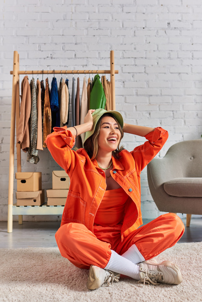 overjoyed asiático dressmaker en panama sombrero y brillante ropa sentado en piso en personal atelier - Foto, imagen