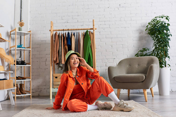 glückliche asiatische Stylistin in orangefarbener Kleidung und Panamahut sitzt auf dem Boden im Atelier, Kleinunternehmen - Foto, Bild