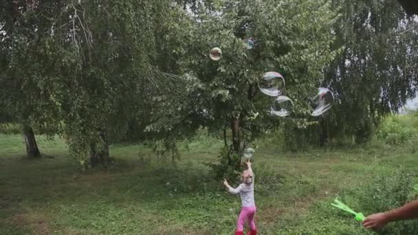 Boldog kislány játszik szappanbuborékokkal a szabadban. Vidám baba nevet, ugrál, buborékokat kap, amit anya fán fúj a gyepen. Vicces gyerek fog kezek buborék fúvó a természetben - Felvétel, videó