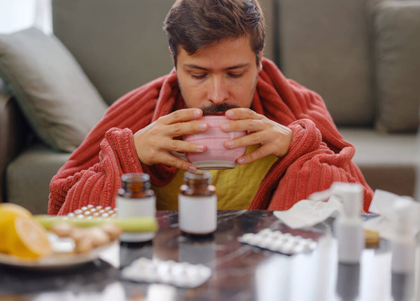 Young Caucasian man with flu on sofa at home. Young Caucasian man with flu feels cold in living room at home drink hot tea in cup. in front of him table are traditional and alternative medicine - 写真・画像