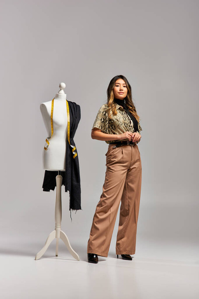 уверенный азиатский стилист смотрит на камеру рядом с манекеном с измерительной лентой и тканью на сером - Фото, изображение