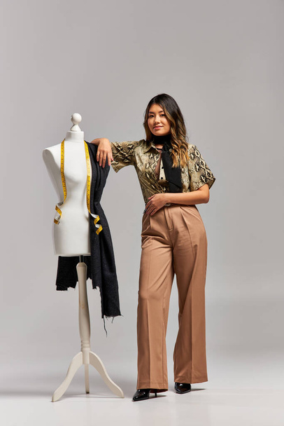 onnellinen trendikäs aasialainen stylisti poseeraa lähellä mallinukke kangas ja mittanauha harmaa tausta - Valokuva, kuva