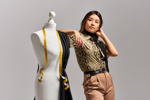 творческий азиатский дизайнер смотрит в сторону рядом с манекеном с тканью и измерительной лентой на сером - Фото, изображение