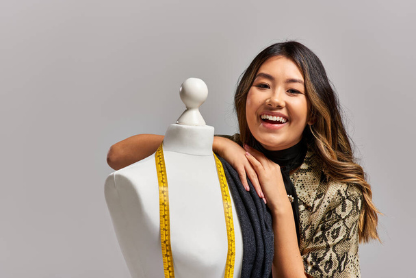 vzrušený asijský stylista se usmívá na kameru poblíž figuríny s tkaninou a měřicí páskou na šedé - Fotografie, Obrázek