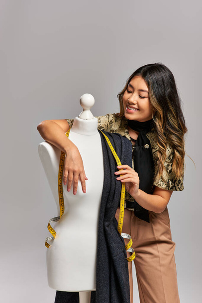 довольный азиатский дизайнер позирует рядом с манекеном с тканью и измерительной лентой на сером фоне - Фото, изображение