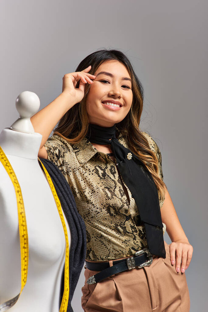 sonriente asiático diseñador de moda con cinta métrica cerca maniquí con tela en gris telón de fondo - Foto, imagen