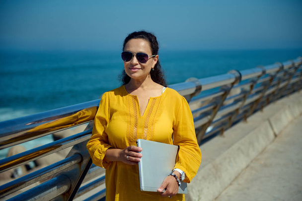 Впевнена красива латиноамериканка в сонцезахисних окулярах, тримає ноутбук і дивиться на камеру, стоїть на тлі Атлантичного океану на міській набережній. Люди. Стиль життя. Віддалена робота - Фото, зображення