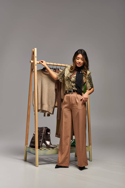 модний азіатський дизайнер одягу, що стоїть біля стійки з модним одягом на сірому фоні - Фото, зображення