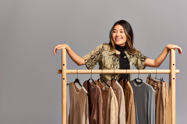 talentierte asiatische Modedesignerin lächelt in der Nähe von Regal mit maßgeschneiderten Kleidern vor grauem Hintergrund - Foto, Bild
