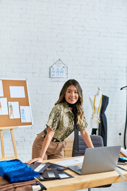 веселый азиатский персональный стилист смотрит в камеру на рабочем столе с ноутбуком в ателье, малого бизнеса - Фото, изображение