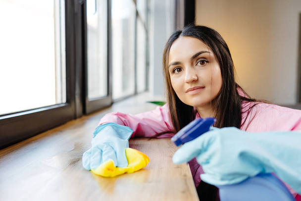 Frau in Schutzhandschuhen lächelt und wischt Staub mit Spray und Staubwedel ab, während sie ihr Haus putzt, Nahaufnahme. - Foto, Bild