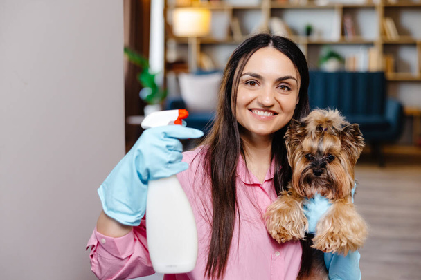 Νεαρή γυναίκα ακουμπισμένη στο τραπέζι κοντά σε προϊόντα καθαρισμού στο σπίτι και το σκυλάκι της στέκεται δίπλα της.. - Φωτογραφία, εικόνα