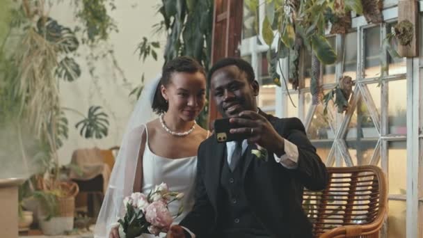 Medio colpo di gioiosa giovane coppia afroamericana seduta su panchina di vimini in camera piena di piante tropicali che scattano ritratti di selfie su smartphone dopo la cerimonia nuziale - Filmati, video