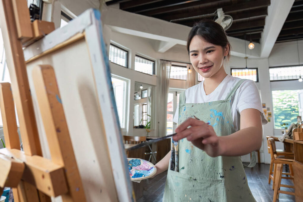 zbliżenie zdjęcie szczęśliwej młodej Azjatki malującej pędzel na płótnie w pracowni malarskiej. Na warsztatach plastycznych.. - Zdjęcie, obraz