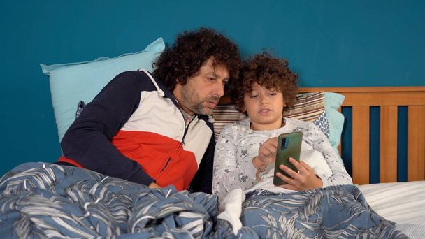Baba ve oğul çocuk 9 yaşında. Uykuya dalmadan önce yatakta internet üzerinden akıllı telefon izliyorlar. Kötü alışkanlık ve çocuklukta internet bağımlılığı. - Fotoğraf, Görsel