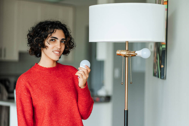 Mujer sonriente de Oriente Medio sosteniendo la bombilla para la lámpara de araña y mirando a la cámara en casa. Chica cambia la bombilla quemada con el fin de lograr una iluminación de alta calidad en la casa - Foto, imagen