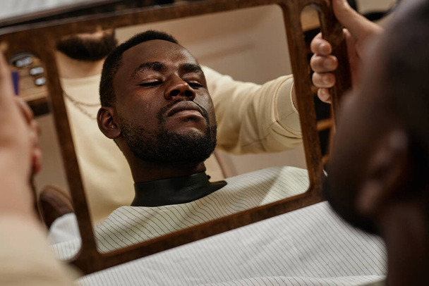 Αφροαμερικάνος πελάτης κοιτάζει τον καθρέφτη και θαυμάζει την περιποιημένη γενειάδα του στο κουρείο. - Φωτογραφία, εικόνα