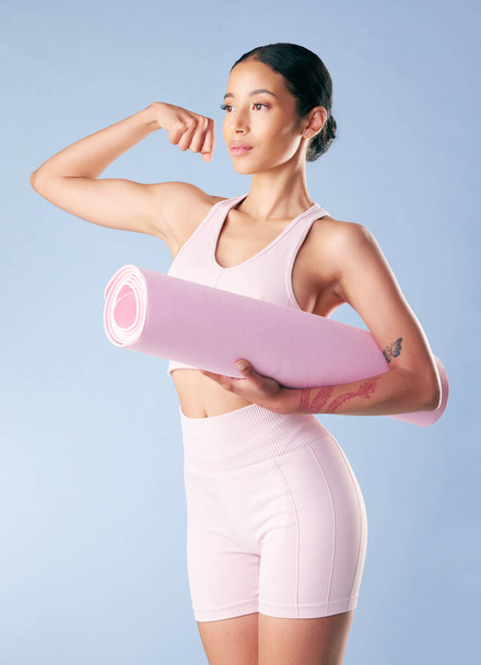 Mixed race fitness woman standing with her yoga mat and flexing her bicep in studio against a blue background. Beautiful young hispanic female athlete exercising or working out. Health and fitness. - Foto, Imagem