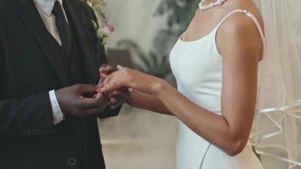 Cropped shot of unrecognizable Black groom in classic black suit putting on wedding ring on finger of his beloved bride during traditional ceremony indoors - Footage, Video