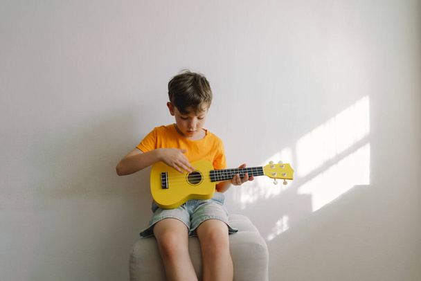 Милый мальчик учится играть на жёлтой гитаре Укулеле дома. Уютный дом. Летние каникулы. - Фото, изображение