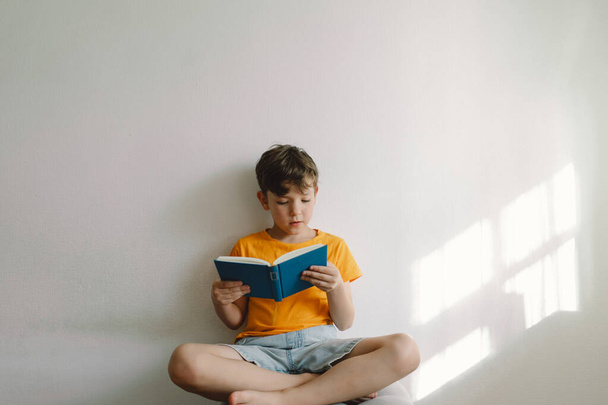 Um menino bonito vestindo uma camiseta laranja está sentado em um otomano macio lendo um livro. Espaço para cópia. Pessoas reais e estilo de vida - Foto, Imagem