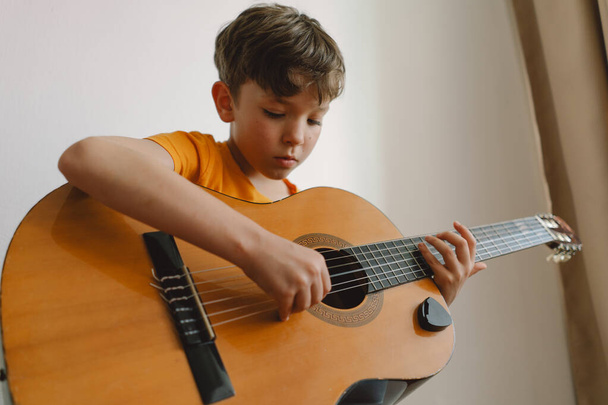 Lindo chico aprende a tocar la guitarra clásica en casa. Acogedora casa. Vacaciones de verano estilo. - Foto, imagen