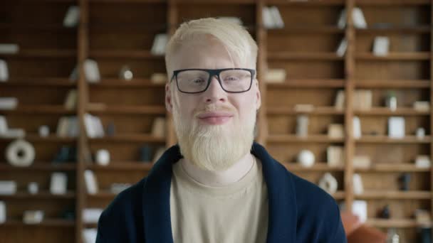 Zbliżenie albinosem ze szczęśliwą twarzą stojącym w bibliotece. Młody biznesmen uśmiechnięty do kamery. Pozytywny prezes w garniturze i okularach w biurze. Portret mądrali w środku. Nietypowy wygląd  - Materiał filmowy, wideo