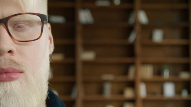 Albino guy with half face posing at camera with book shelves on the background. Young businessman wearing fashionable glasses. Closeup portrait of smiling man. Hipster with happy emotion, copy space - Footage, Video