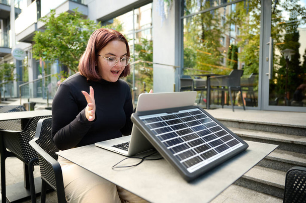 Frauen nutzen tragbare Solarzellen, um Laptop und Smartphone aufzuladen. Porträt einer jungen Studentin, die ferngesteuert vom Laptop an einem Tisch in einem Café sitzt. Überraschte Frau. Erneuerbare Energien. - Foto, Bild