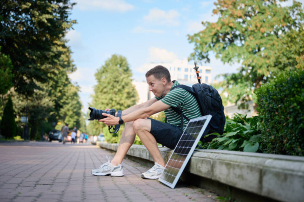 Fotógrafo viajante usando painel solar para carregar sua câmera. Jovem viajante esperando por sua câmera para carregar. Relaxamento masculino positivo após longa caminhada. - Foto, Imagem